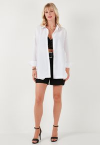 Witte oversized button-up shirt met lange mouwen, gecombineerd met zwarte shorts en een zwarte kanttop. Model draagt zwarte haksandalen.