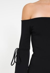 Svart off-shoulder topp med långa ärmar, som har knutdetaljer vid ärmsluten och ett mjukt, stretchigt tyg.