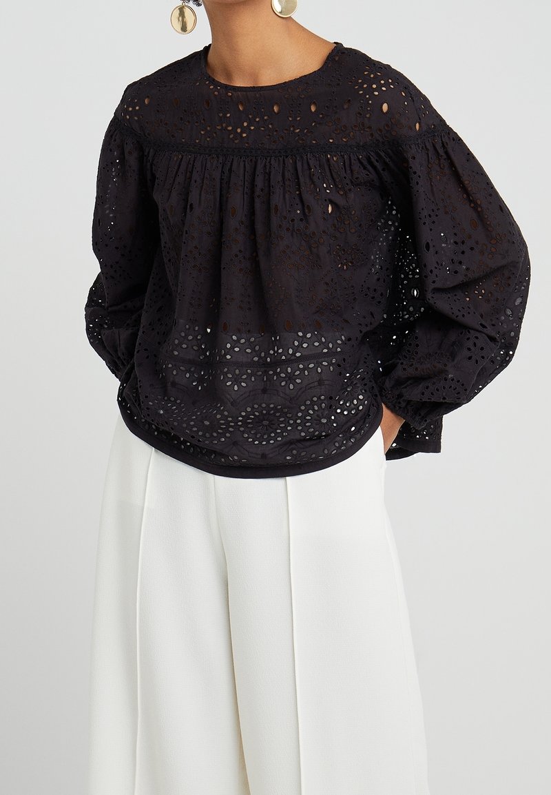 Blouse noire en œillets avec un col rond, des manches bouffantes et des motifs découpés complexes. Portée sur un pantalon large blanc.