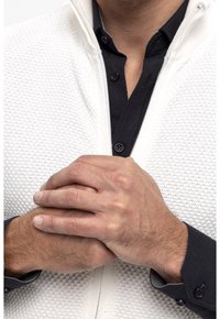 Chaqueta blanca texturizada con cremallera, llevada sobre una camisa negra abotonada, con las manos entrelazadas al frente, mostrando el contraste de material y color.