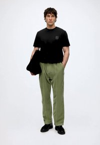 Homme portant un t-shirt noir, pantalon vert olive, chaussures noires, tenant un sac noir avec une main dans la poche, sur un fond clair et uni.