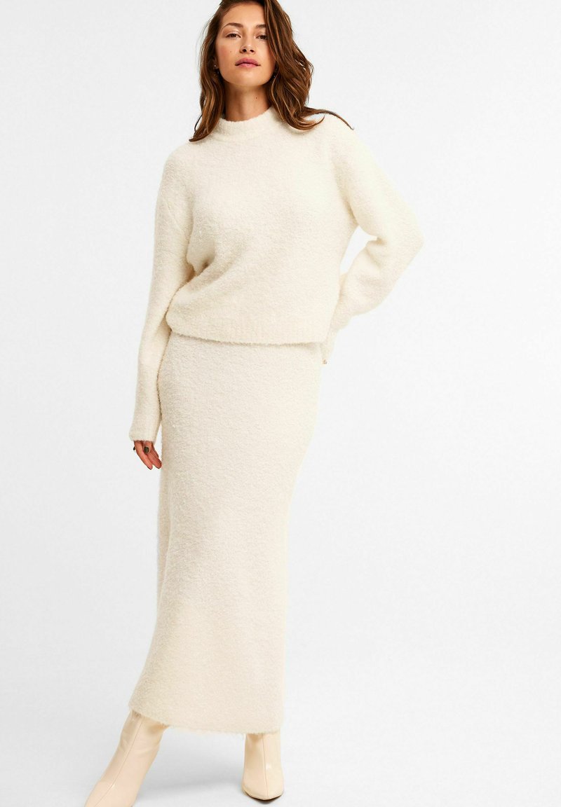 Creme strikket sweater og matchende gulvlængde nederdel, begge med en blød tekstur. Outfitet har høj hals og lange ærmer.