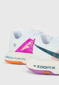 Chaussures de course blanches Nike montrant un gros plan des talons avec logos roses et noirs, marquage Flyplate et ZoomX, et semelles orange et violettes.