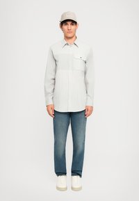 Bărbat îmbrăcat cu o cămașă ușor gri cu modele, blugi, adidași albi și o pălărie bej, stând în fața unui fundal alb simplu.