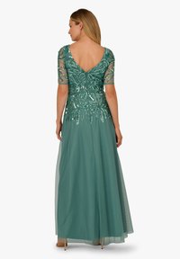 Kobieta w turkusowej sukni z cekinowymi kwiatowymi wzorami na przezroczystych półrękawach i górnej części pleców, stojąca tyłem z rozpuszczonym blond włosami.