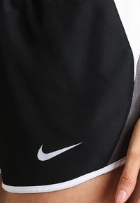 Mustat urheilushortsit, joissa on valkoinen Nike-merkki vasemmalla jalalla. Henkilöllä on käsi lepäämässä helman lähellä.