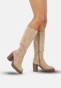Botas de ante marrón claro hasta la rodilla con un tacón de madera grueso y suela de goma. Presentan un cierre de cremallera lateral y detalles de costura sutiles.