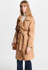 Manteau trench en faux suède beige avec une taille ceinturée, des épaulettes et des détails de boutons. Associé à un jean foncé à pattes d'éléphant et un petit sac à main.