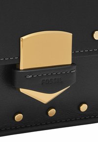 Sac en cuir noir avec une fermeture à clasp doré, logo embossé et clous dorés sur une texture lisse avec des surpiqûres grises contrastantes.