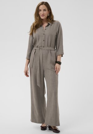 Kvinde med langt hår iført en taupe buksedragt med bælte, knapper og trekvartærmer, stående og smilende mod en ensfarvet baggrund.