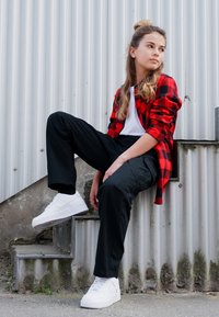 Chemise à carreaux rouge et noire, t-shirt blanc, pantalon cargo noir et baskets blanches. Assis sur des escaliers contre un fond en métal ondulé.