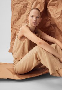 Junge Frau mit zurückgebundenem Haar, die ein beiges ärmelloses Top und Hosen trägt, sitzt vor einem zerknitterten braunen Papierhintergrund und schaut in die Kamera.