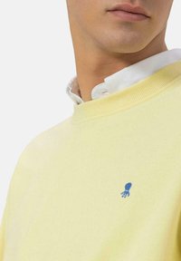 Sudadera amarilla con cuello acanalado, presenta un pequeño logo de un pulpo azul en el pecho. La tela parece suave y texturizada, sobre un fondo blanco.