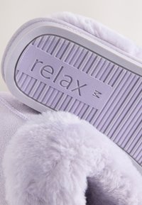 Chausson doux lavande avec bordure en fourrure duveteuse, montrant la semelle avec adhérence texturée et le mot « relax » embossé en dessous.