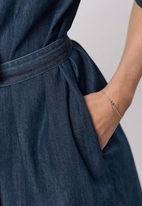 Tmavě modré džínové šaty s vázáním v pase, zdobené sklady a boční kapsou. Na zápěstí zdobí stříbrný náramek s jemným řetízkovým detailem.