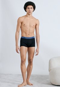 Μαύρα ανδρικά εσώρουχα boxer με μπλε ζώνη μέσης, που φέρουν λογότυπο. Το μοντέλο στέκεται με φυσική στάση μπροστά σε ανοιχτόχρωμο φόντο.