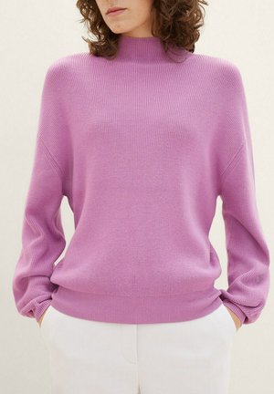 Femme portant un pull côtelé violet clair à col montant et manches longues, associé à un pantalon blanc, les mains dans les poches.