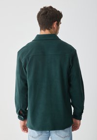 Maglia in fleece verde scuro con colletto, polsini bottonati e vestibilità rilassata. Superficie testurizzata con un design classico.