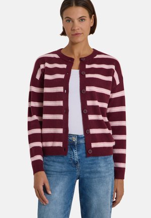 Femme portant un cardigan boutonné rayé bordeaux et rose, un haut blanc en dessous et un jean bleu, debout devant un fond uni.