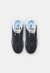 Črne semiš in mrežaste teniske z belimi detajli, črnimi vezalkami in rjavim gumijastim podplatom. Vsebujejo blagovno znamko in oznako nasmeška na jeziku.