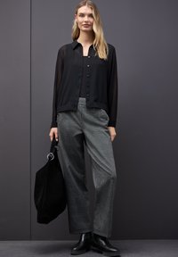 Sort gennemsigtig skjorte med knapper over en sort tanktop, parret med tjekkede grå brede bukser. Modellen holder en sort tote bag og bærer sorte støvler.