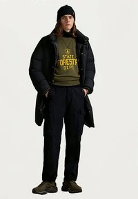 Młoda osoba w czapce beanie, czarnej kurtce puchowej, zielonej bluzie z napisem "Departament Leśnictwa Stanu", czarnych spodniach cargo i brązowych sneakerach, stoi z rękami w kieszeniach.