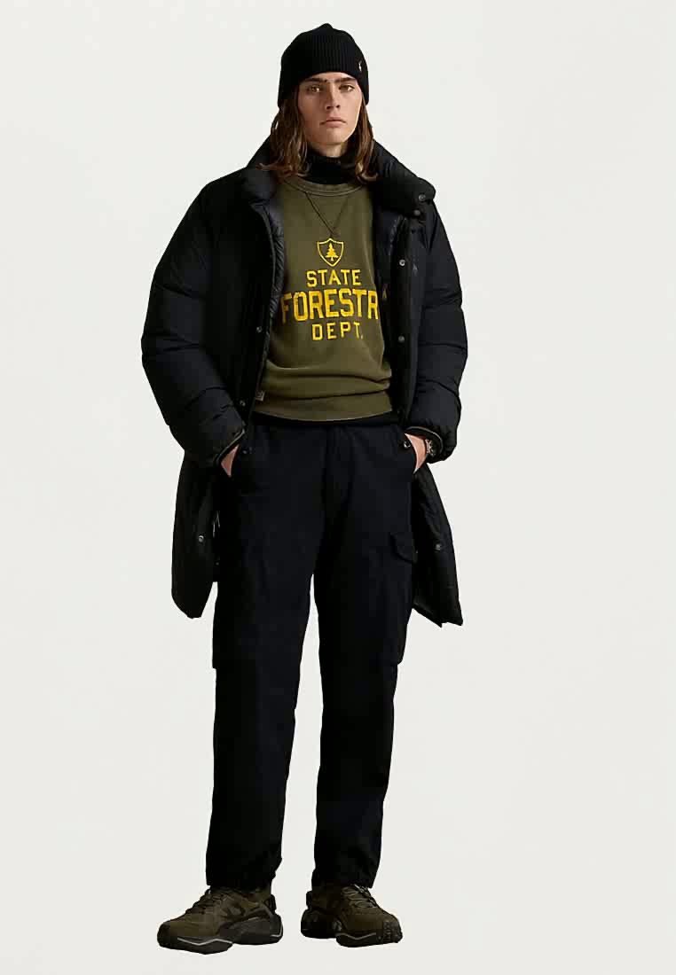 Młoda osoba w czapce beanie, czarnej kurtce puchowej, zielonej bluzie z napisem "Departament Leśnictwa Stanu", czarnych spodniach cargo i brązowych sneakerach, stoi z rękami w kieszeniach.