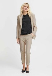 Blazer beige con una vestibilità su misura, abbinato a pantaloni tapered beige e una t-shirt nera. Il modello indossa ballerine nere, con capelli disordinati.