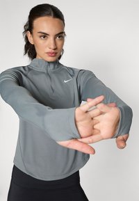 Lys grå sportslig langermet topp med kvart glidelås og tommelhull-ermer, med en liten hvit logo på brystet. Glatt tekstur.