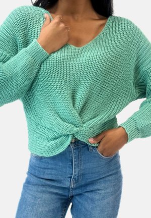 Kvinde iført en mintgrøn strikket sweater med knude foran og blå high-waisted jeans, hånd hvilende ved nakken og lommen.