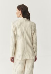 Beige pruhovaný blazer s přiléhavým střihem, zdobený vertikálními černými proužky a klopou. Tkanina vypadá hladce a lehce.