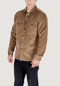Camicia in corduroy marrone con chiusura a bottoni sul davanti, due tasche sul petto e taglio dritto. Tessuto testurizzato e dettagli in bottoni neri visibili.