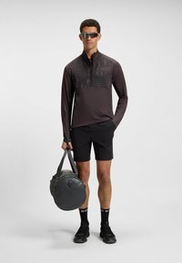 Sportig långärmad topp i mörkgrå med ett mönstrat bröst, parat med svarta shorts, svarta sneakers och en grå gymbag.