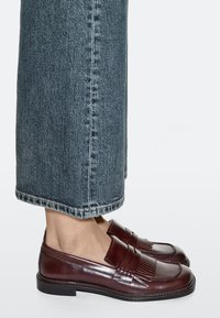Brown leather loafers with fringe detailing, flat sole, and structured design, paired with wide-leg denim jeans, rolled at the cuff.
