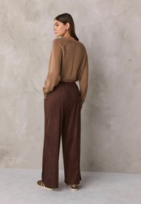 Brauner Strickpullover mit langen Ärmeln, Rückennaht-Detail, kombiniert mit dunkelbraunen, weit geschnittenen Hosen. Schuhe sind goldakzentuiert. Einfarbiger Hintergrund.