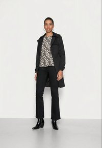 Casaco preto de trench sobre uma camisa com padrão floral, combinado com calças largas pretas e botas de salto alto. A modelo está de pé sobre um chão liso de cor clara.
