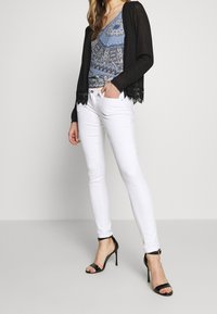 Svart cardigan med spetsdetalj över en blåmönstrad topp, kombinerad med vita tajta jeans och svarta högklackade sandaler.