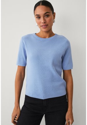Kvinde med mørkt hår iført en lyseblå kortærmet sweater og sorte bukser står mod en ensartet lys baggrund og har sølvfarvede creol-øreringe på.