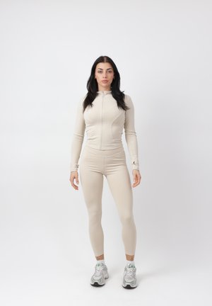 Frau mit langen dunklen Haaren steht mit Blick nach vorn und trägt eine beige, eng anliegende Jacke mit Reißverschluss, passende Leggings und weiße Turnschuhe vor weißem Hintergrund.