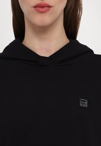 Svart hettegenser i ribbestrikket stoff, med en liten rektangulær logo-patch på venstre side. Glatt tekstur, avslappet passform.