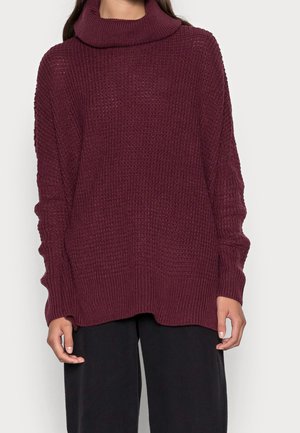 Femme portant un pull à col roulé en maille gaufrée bordeaux oversize avec manches longues et pantalon ample noir sur fond uni.