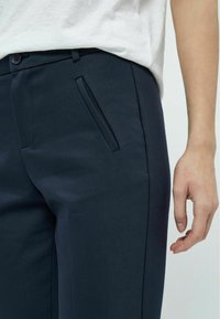Pantaloni blu navy con una texture liscia, dotati di una tasca laterale e una chiusura con bottone, cuciture a contrasto e una vestibilità su misura.