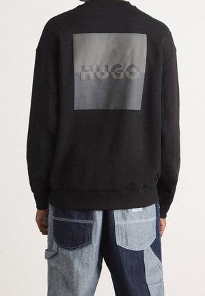 Person trägt einen schwarzen Sweatshirt mit einem grauen Quadrat und dem Schriftzug "HUGO" auf der Rückseite, kombiniert mit zweifarbigen Patchwork-Jeans.