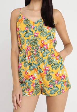 Femme portant une combinaison jaune sans manches avec des feuilles tropicales vertes et des fleurs roses, avec une taille cintrée et des poches.