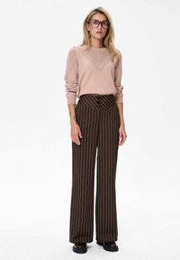Vrouw met grote bril, ringen in de oren, een roze gebreide trui, bruine gestreepte wijde broek met hoge taille en zwarte schoenen staat.