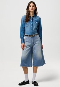Jeansskjorta i medelblått med spetsig tass, två bröstfickor och knappar. Vida ljusblå jeansculotter med blekt mönster. Svart bälte.