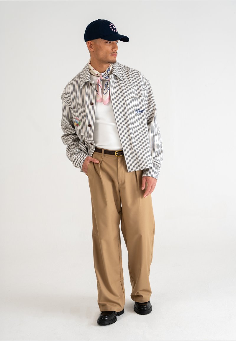 Gestreepte button-up jacket, witte T-shirt, beige broek, marineblauwe pet en een roze patroon sjaal. Zwarte schoenen. Effen kleur achtergrond.