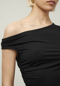 Musta olkaimeton paita, jossa on rypytetty yksityiskohta sivussa, valmistettu pehmeästä ja joustavasta kankaasta. Ominaisuuksiin kuuluu sileä pinta ja istuva muotoilu.