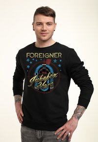 Felpa nera con grafica di un jukebox e testo colorato "FOREIGNER Jukebox Hero", maniche lunghe e scollo a girocollo.