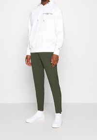 Felpa bianca con cappuccio e lacci, logo sul petto, abbinata a pantaloni a sigaretta verde oliva e sneakers bianche alte.